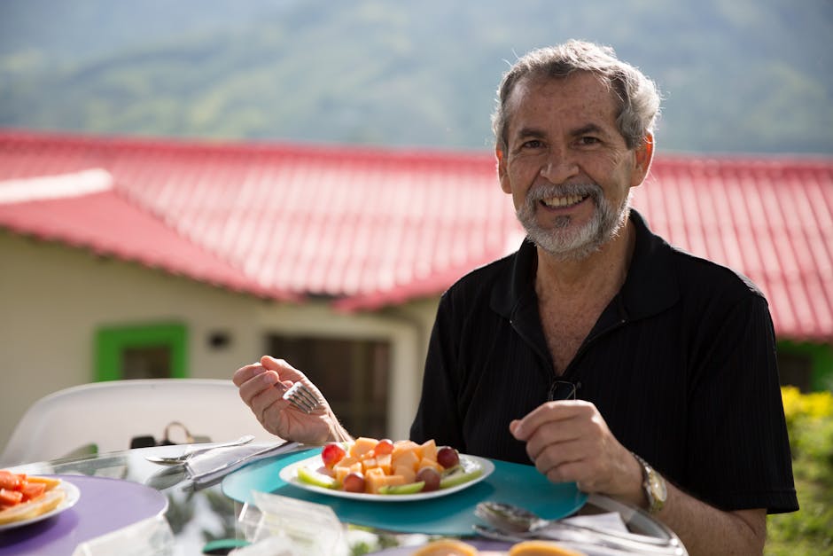 A joyful senior man with gray hair enjoying a healthy fruit meal at a table outdoors, with a scenic background.