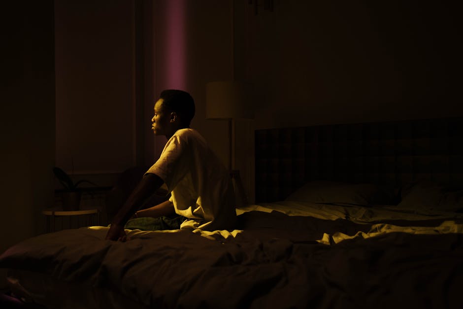 A person in deep thought sitting on a bed in a dimly lit bedroom, creating an introspective mood.