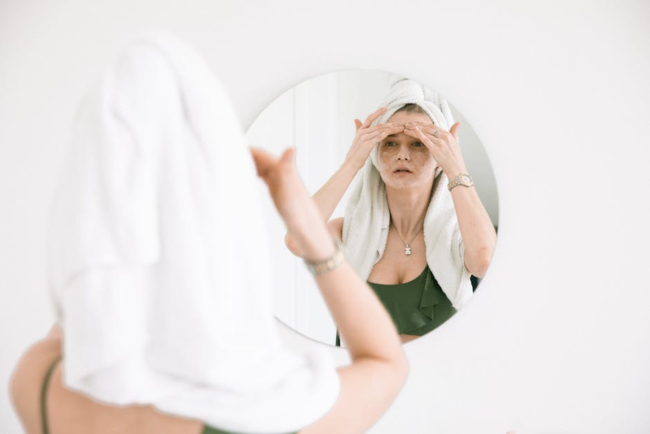 Woman with towel on head applies skincare, reflects self-care routine in mirror.