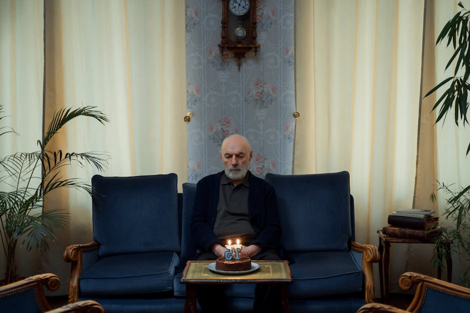 An elderly man sits alone with a lit birthday cake indoors, suggesting contemplation and solitude.