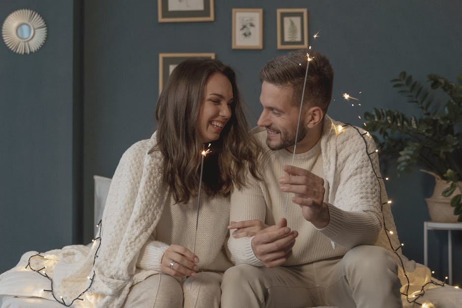 A loving couple smiles warmly while playing with sparklers in a cozy bedroom setting.